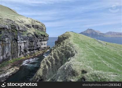 Horizontal scenery image to the sea-filled gorge nearby idyllic village Gjogv, most northern village on the island of Eysturoy in foreground surrounded by high cliffs. Glorious sceneries of the Faroes. Postcard motif.