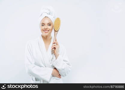 Horizontal shof of charming beauty woman has broad smile, perfect white teeth, dressed in bath robe, holds anti cellulite massage brush, poses against white backgruound, blank space for your promo