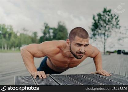 Horizontal shot of active bearded European man does exercises for shouldrs, chest and biceps, stays in good physical shape, poses outdoor, has muscled arms. Sport, workout and push up concept