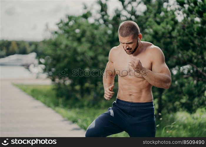 Horizontal shot of active bearded man has active exercises outdoor runs on one place has naked torso muscular body listens music in earphones poses against green trees. Sport and motivation concept