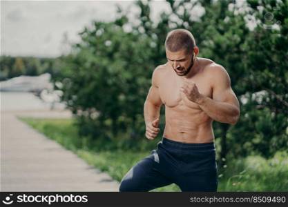 Horizontal shot of active bearded man has active exercises outdoor runs on one place has naked torso muscular body listens music in earphones poses against green trees. Sport and motivation concept