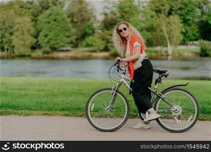 Horizontal shot of active female bicyclist wears t shirt, black pants and sneakers, stops near river side, keeps feet on pedals of bicycle beautiful scenic nature in background. Sunny day, active rest