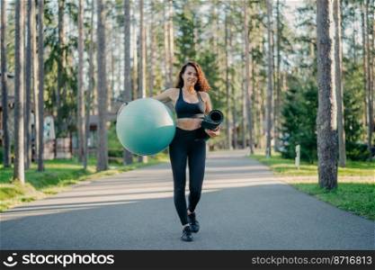 Horizontal shot of active slim woman dressed in sportswear, carries fitball and rolled up karemat, walks on road near trees, going to have workout, breathes fresh air, leads healthy sporty lifestyle