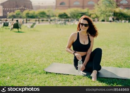 Horizontal shot of active slim woman in sportswear sits on karemat holds bottle of cold water has rest after exercising abdominal muscles poses on green lawn wears sunglasses. Workout at street