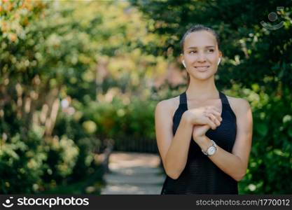 Horizontal shot of active sporty woman with dark hair healthy skin keeps hands together prepares for training focused into distance smiles happily enjoys nature and fresh air. People sport lifestyle