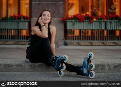 Horizontal shot of active young woman takes break while riding rollers at street dressed in black sportsclothes feels satisfied smiles pleasantly poses outdoor. Summer lifestyle and rest concept