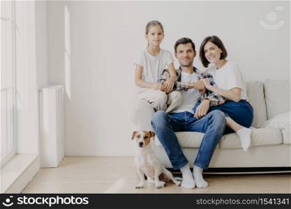 Horizontal shot of affectionate family pose together on couch in empty spacious room with white walls, their favourite dog sits on floor. Copy space aside. Happy female child glad be with mom and dad