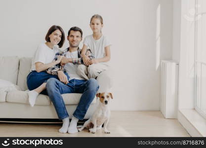 Horizontal shot of affectionate family pose together on couch in empty spacious room with white walls, their favourite dog sits on floor. Copy space aside. Happy female child glad be with mom and dad