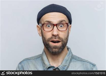 Horizontal shot of amazed excited shocked man stares at camera with unexpected expression, keeps mouth opened, hears awful news, isolated over grey background. Omg and great surprisment concept