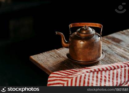 Horizontal shot of antique kettle on vintage wooden table. Old crockery. Metal copper teapot against dark background red towel near. Selective focus