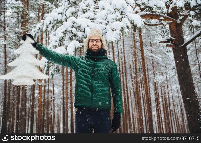 Horizontal shot of attractive bearded guy stands against winter trees in forest, holds white artificial fir tree, expresses festive mood and preapres for winter holidays. People, weather, season