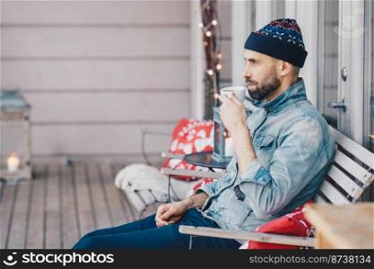 Horizontal shot of attractive bearded male wears hat and denim jacket, drinks hot tea, sits at bench on balcony, has thoughtful expression, thinks about something important in life. Leisure time