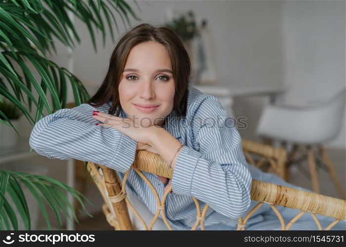 Horizontal shot of attractive calm young Caucasian woman has pleased facial expression, enjoys domestic atmosphere, poses at rattan furniture, near green plant. People, beauty and rest concept