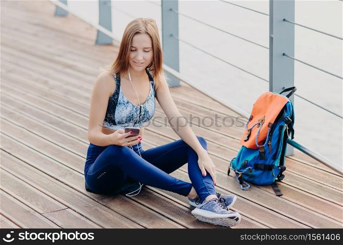 Horizontal shot of attractive Caucasian woman feels relaxed, sits crossed legs, looks at smart phone, types text message, listens favourite track in earphones. Peoople, online communication concept