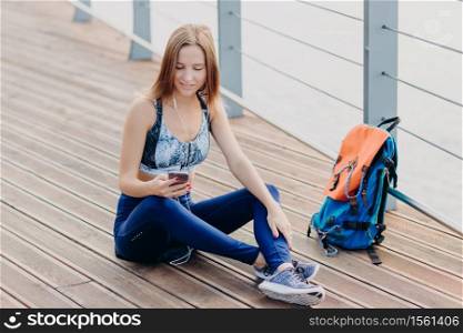 Horizontal shot of attractive Caucasian woman feels relaxed, sits crossed legs, looks at smart phone, types text message, listens favourite track in earphones. Peoople, online communication concept