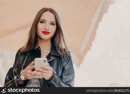 Horizontal shot of attractive dark haired female with red painted lips, dressed in black leather jacket, holds modern cell phone, sends text messages, chats online, strolls at city street. Technology