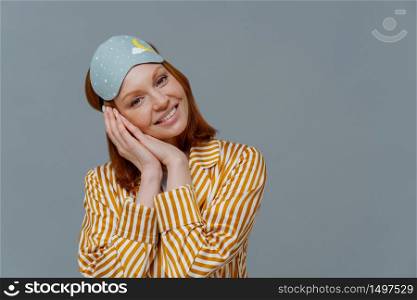 Horizontal shot of attractive ginger woman feels glad after having enough sleep, just woke up, tilts head on hands, wears blindfold and pajamas, isolated on grey wall, empty space. Sleep gesture