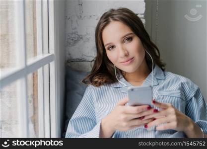 Horizontal shot of attractive girl enjoys radio broadcasting via cell phone and earphones, connected to high speed internet, entertains herself with listening music, poses on window sill at home