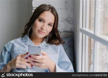 Horizontal shot of attractive girl enjoys radio broadcasting via cell phone and earphones, connected to high speed internet, entertains herself with listening music, poses on window sill at home