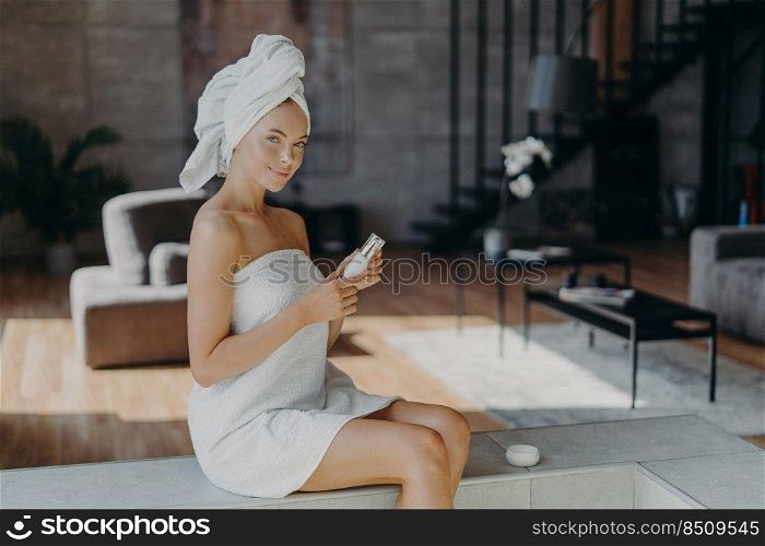 Horizontal shot of attractive lovely woman sits against cozy room background applies body lotion after taking bath uses cosmetic product for glowing skin wrapped in towel undergoes beauty treatments
