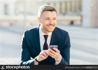 Horizontal shot of attractive male with cheerful thoughtful expression, uses modern mobile phone for searching information on internat website, connected to high speed 4G internet, makes money online