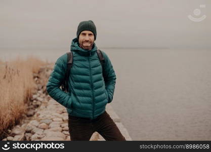 Horizontal shot of attractive unshaven man has cheerful expression, wears hat and jacket, keeps hands in pocket, carries rucksack, has walk during foggy autumn weather, poses near lake outdoor