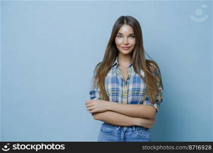 Horizontal shot of attractive young female with long straight hair, wears checkered shirt, jeans, keeps hands crossed, has clean skin, minimal makeup, expresses positive emotions, enjoys photo session