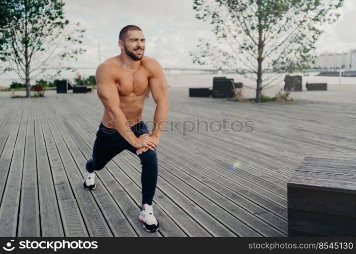 Horizontal shot of bearded motivated man stretches leg muscle before sport exercises or running, does warm up, poses at city park outdoor, has muscular torso. Sportsman goes in for sport regularly