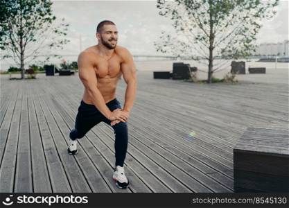 Horizontal shot of bearded motivated man stretches leg muscle before sport exercises or running, does warm up, poses at city park outdoor, has muscular torso. Sportsman goes in for sport regularly