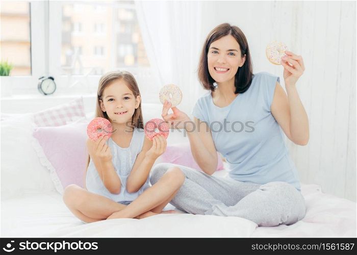 Horizontal shot of beautiful brunette femle with bobbed hairstyle, dressed in pyjamas, sits near her small daughter, hold together doughnuts, going to have breakfast in bed, has pleasant smiles