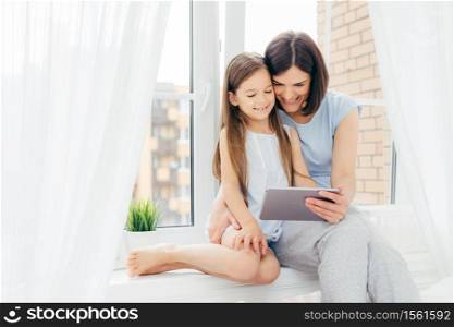 Horizontal shot of beautiful brunette mother and daughter spend free time together, sit on window sill, watch interesting movie via digital tablet, being in bedroom. People, technology, family concept