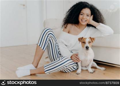 Horizontal shot of beautiful curly woman leans at sofa, sits on floor near pedigree dog, wears sweater, striped pants and socks. African American lady poses with pet. People and animals concept