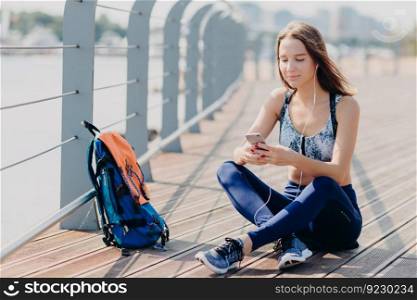 Horizontal shot of beautiful happy atthletic female in sneakers, sportswear, feels relaxed while sits outdoor, uses modern cell phone for surfing internet, dowloads music for playlist, listens song