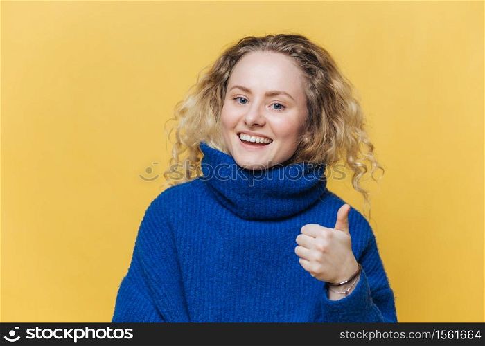 Horizontal shot of beautiful smiling female in casual sweater keeps thumb raised, demonstrates her agreement and approval, poses against yellow studio background. Happy young woman says: Yes