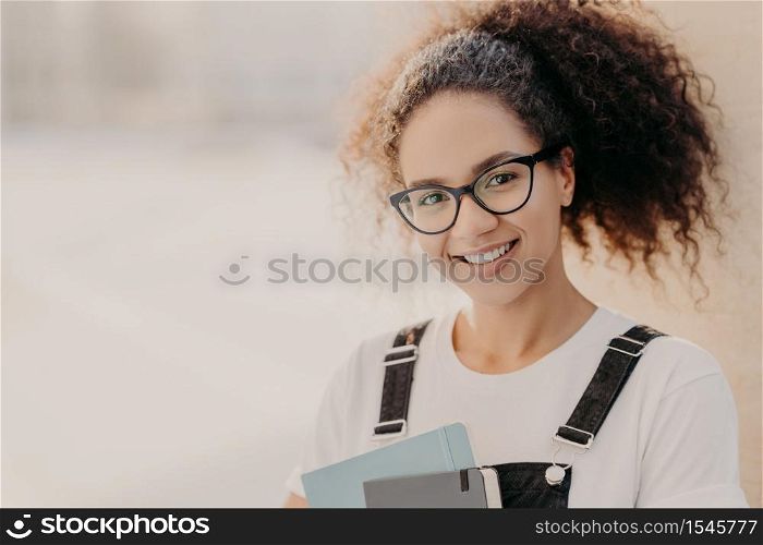 Horizontal shot of beautiful woman with crisp hair combed in pony tail, carries notepad, wears white t shirt and overalls, enjoys studying, poses against blurred background with empty space for text