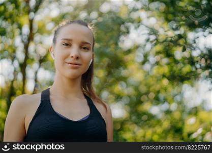 Horizontal shot of beautiful young woman with dark combed hair enjoys outdoor workout listens music in wireless earphones poses against green blurred nature background. Sport fitness healthy lifestyle