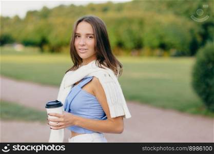 Horizontal shot of brunette lady with slim body, dressed in t shirt, white sweater on shoulders, drinks aromatic coffee, stands outdoor against green nature background. People and lifestyle concept