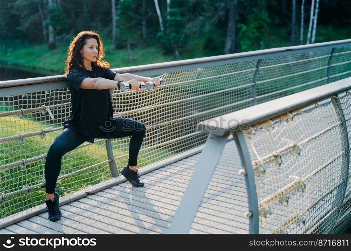 Horizontal shot of brunette woman excersises with dumbbells, does squats, dressed in active wear, poses at bridge, being motivated, has workout exercises, leads sporty lifestyle. Fitness, workout