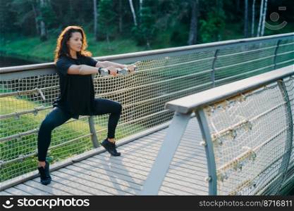 Horizontal shot of brunette woman excersises with dumbbells, does squats, dressed in active wear, poses at bridge, being motivated, has workout exercises, leads sporty lifestyle. Fitness, workout