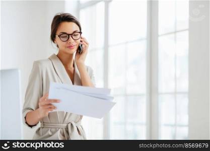Horizontal shot of business lady discusses details of contract, works in office, concentrated on information from accountings, holds paper documents during telephone conversation, dressed formally
