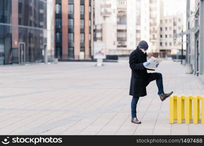 Horizontal shot of businessman stands outdoor near office building, reads newspaper, wears medical mask and gloves during quarantine, prevents coronavirus. Pandemic situation in whole world.