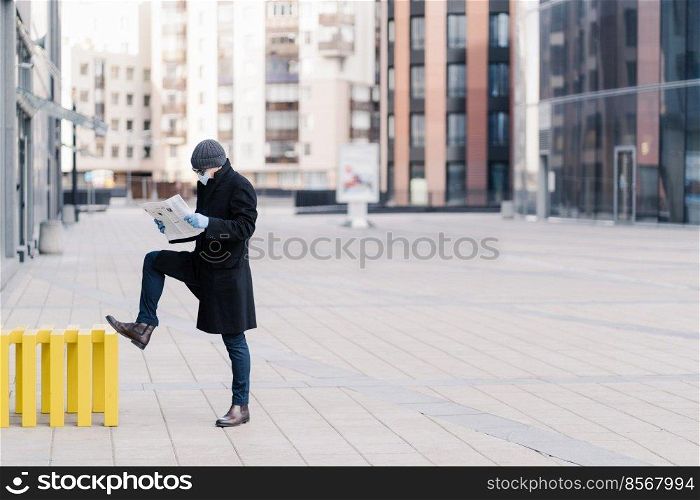 Horizontal shot of businessman stands outdoor near office building, reads newspaper, wears medical mask and gloves during quarantine, prevents coronavirus. Pandemic situation in whole world.