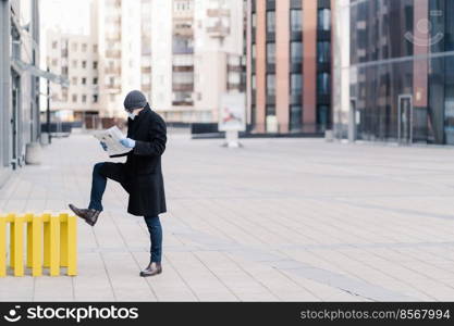 Horizontal shot of businessman stands outdoor near office building, reads newspaper, wears medical mask and gloves during quarantine, prevents coronavirus. Pandemic situation in whole world.