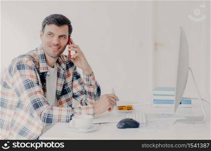 Horizontal shot of busy male freelancer has telephone conversation, dressed in checkered shirt, writes down information, sits at desktop with computer, solves financial problem, poses at workplace