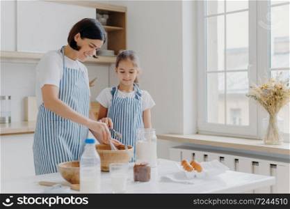 Horizontal shot of caring mother and daughter cook together, mom gives culinary lesson to small girl, use flour, eggs and milk to prepare tasty meal. Family time, baking and culinary concept