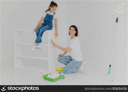 Horizontal shot of caring mother and her daughter busy refurbishing furniture at home, use white paint and paintbrush, roller, happy almost finishing work, busy painting new house, dressed casually