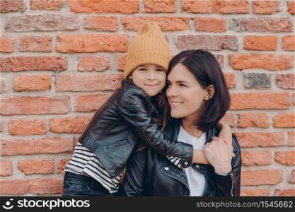 Horizontal shot of charming friendly little child and her mother embrace each other, dressed in black leather jackets, have appealing look and pleasant smile pose over brick wall. Relationship concept