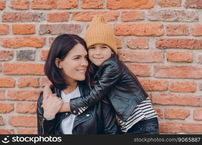 Horizontal shot of charming friendly little child and her mother embrace each other, dressed in black leather jackets, have appealing look and pleasant smile pose over brick wall. Relationship concept