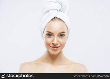 Horizontal shot of charming tender Euoropean woman with healthy skin after taking bath, enjoys hygienic procedures, has natural makeup, wears soft towel on head, stands bare shoulders indoor