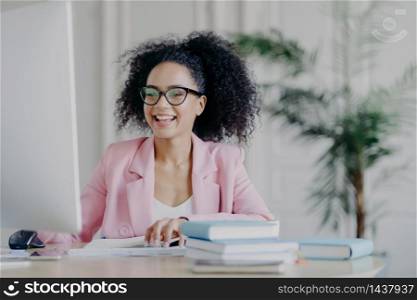 Horizontal shot of cheerful African American freelancer browses internet on computer, focused at screen, wears optical glasses and rosy formal costume, poses at desktop with pile of books, papers